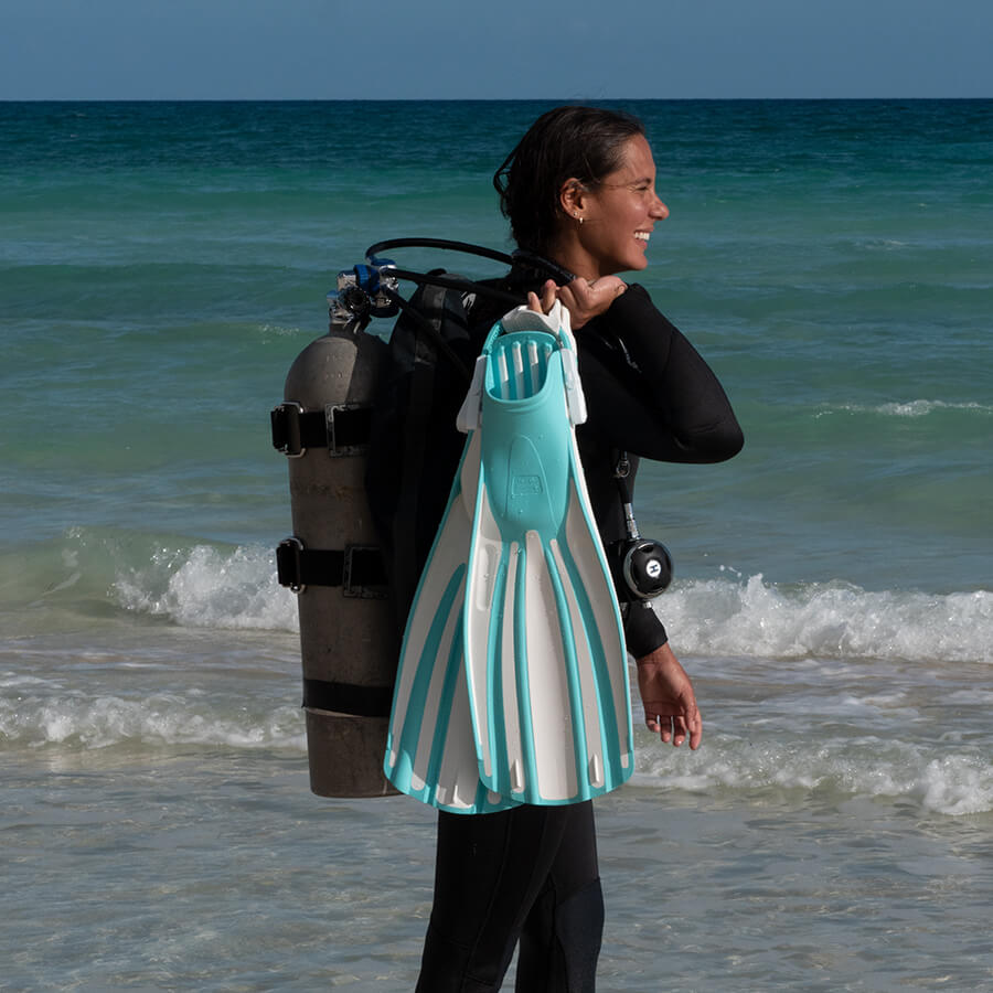female scuba diver walking by the sea with a cylinder on her back carrying a set of blue fins at her side